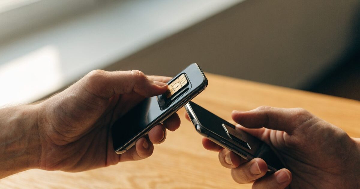 Due mani spostano una scheda SIM dal vassoio di uno smartphone a quello di un altro telefono, sopra una scrivania di legno illuminata da una finestra