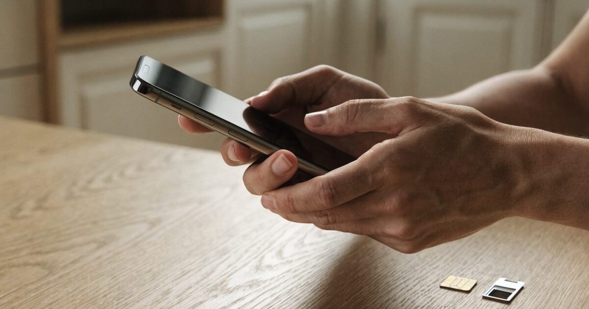Due mani tengono uno smartphone sopra un tavolo di legno chiaro; accanto, appoggiate al piano, una scheda SIM e il vassoio porta-SIM estratto